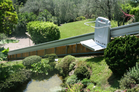 Outdoor platform lift installed on an inclined rail system, providing wheelchair accessibility along a landscaped hillside surrounded by trees and shrubs.