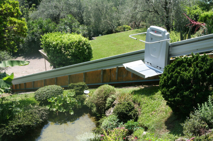 Outdoor platform lift installed on an inclined rail system, providing wheelchair accessibility along a landscaped hillside surrounded by trees and shrubs.