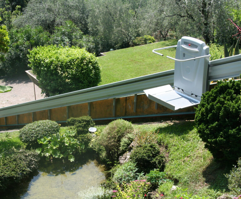 Outdoor platform lift installed on an inclined rail system, providing wheelchair accessibility along a landscaped hillside surrounded by trees and shrubs.