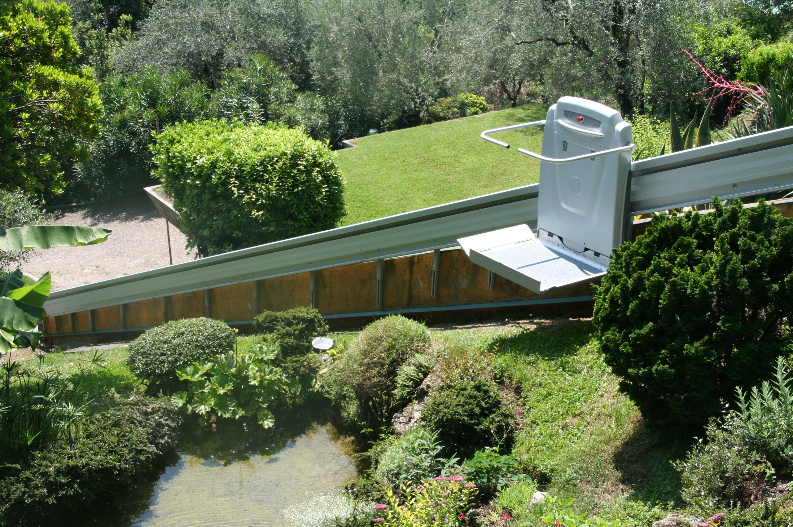 Outdoor platform lift installed on an inclined rail system, providing wheelchair accessibility along a landscaped hillside surrounded by trees and shrubs.