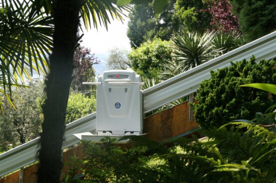 Outdoor platform lift installed on an inclined rail system, providing wheelchair accessibility along a landscaped hillside surrounded by trees and shrubs.