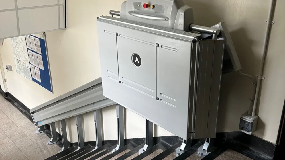 ACCESS BDD inclined platform lift (supra linea) folded in a compact and discrete design on an indoor staircase against a white wall.