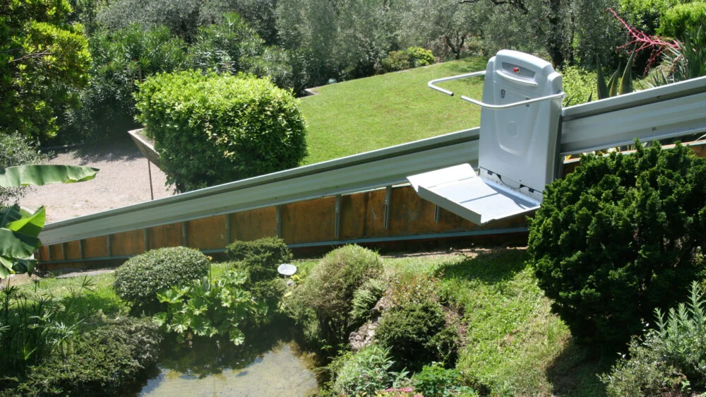 ACCESS BDD inclined platform lift (supra linea) sitting in the middle of the rail outdoors on a hill with a lot of green plant life surrounding it.
