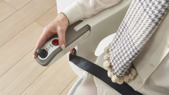 A close-up of a person’s hand operating the joystick and red button on the armrest of a HomeGlide Stair Lift, with the person wearing a houndstooth-patterned scarf and fastened seatbelt.