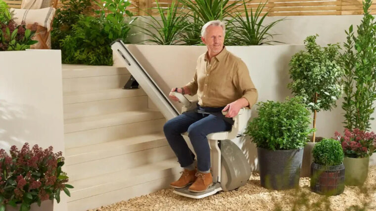An older man sits on a motorized chair lift at the base of outdoor steps, surrounded by green plants and shrubs in a garden setting.