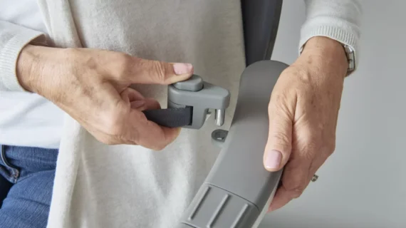 Close-up of a person’s hands assembling a gray metal object—part of the Flow X Curved Stair Lifts—fastening a strap with a small gray plastic latch. The person is wearing a light-colored sweater and a watch.