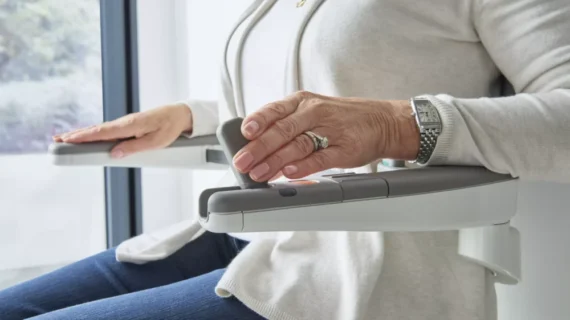 Close-up of an older adult’s hands operating the control of a Flow X Curved Stair Lift, showing a wedding ring and wristwatch, with the person seated in casual clothing near a window.
