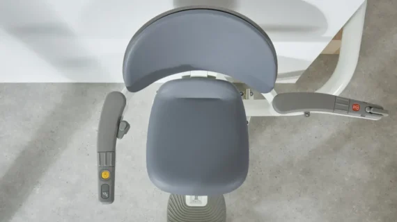 Top-down view of a gray Flow X Curved Stair Lift chair with armrests and control buttons, positioned next to a white wall on a gray floor.