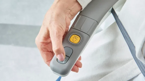 A hand holding a gray remote control device for Flow X Curved Stair Lifts, featuring a yellow WIPER button and two additional buttons—one being pressed. The person is wearing a white garment.