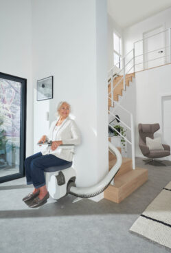 An older woman sits on a Flow X Curved Stair Lift at the base of a modern staircase in a bright, minimalist home, smiling as she prepares to ascend the stairs.