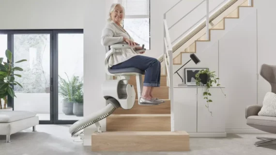 An older woman smiles while riding a Flow X Curved Stair Lift up a wooden staircase in a modern, bright living room with large windows and contemporary furniture.