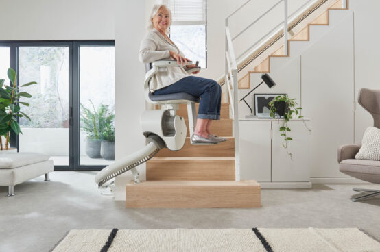 An older woman smiles while riding a Flow X Curved Stair Lift up a wooden staircase in a modern, bright living room with large windows and indoor plants.