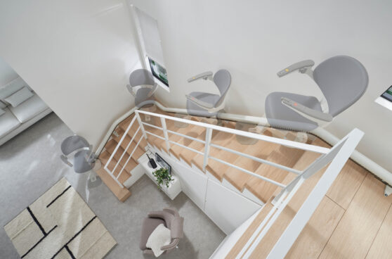 A Flow X Curved Stair Lift with a gray chair is shown moving along a wooden staircase with white railings in a modern, bright home. The image captures multiple positions of the chair ascending the stairs.