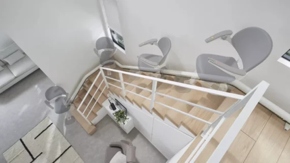 A Flow X Curved Stair Lift with a gray seat is shown gliding along a curved rail beside a modern wooden staircase in a bright, minimalist home. Multiple overlay images illustrate the stairlift’s smooth movement up the stairs.