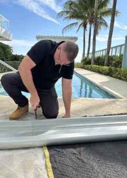 A worker wearing a black shirt and gloves kneels beside a swimming pool, using a tool to install or adjust a long metal platform lift track on the ground outdoors on a sunny day.