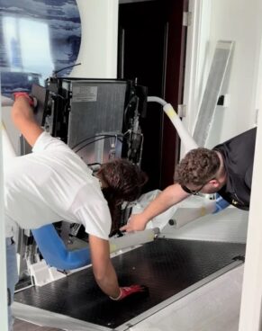 Two technicians are installing and adjusting a platform lift system indoors, leaning over the machinery while working on the base frame and wiring near a doorway.