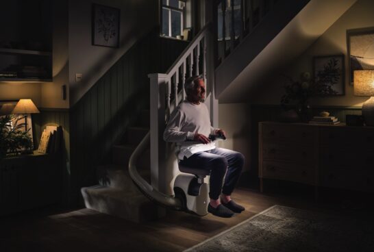 An older man sits on a Flow X Curved Stair Lift at the bottom of a staircase in a dimly lit, cozy living room. Holding a remote control, he appears ready to glide upstairs as the room glows with soft table lamp light.