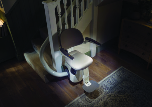 A Flow X Curved Stair Lift with armrests and a footrest is installed along a curved staircase in a home with wooden floors and a patterned rug, positioned at the base of the stairs near the banister.