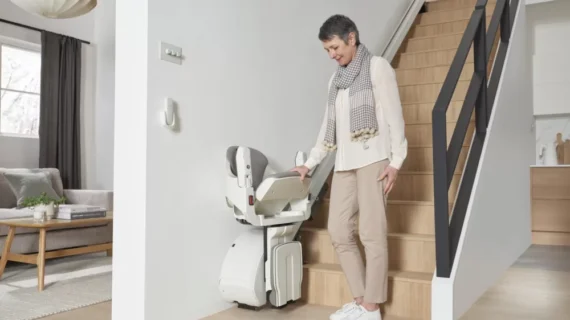A smiling older woman stands beside a HomeGlide Stair Lift at the base of a wooden staircase in a bright, contemporary home.
