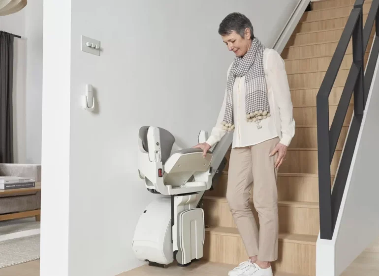 A smiling older woman stands beside a HomeGlide Stair Lift at the base of a wooden staircase in a bright, contemporary home.