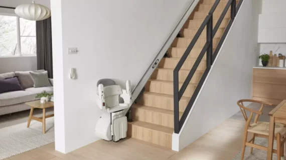 A modern HomeGlide Stair Lift is installed along a wooden staircase in a bright, contemporary home, with a living area on the left and dining area on the right. The stairlift is positioned at the base of the stairs.
