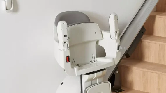 A HomeGlide Stair Lift with a white and gray seat is installed on a wooden staircase, attached to a metal rail along the side. The seat is facing away from the stairs and is in a parked position.