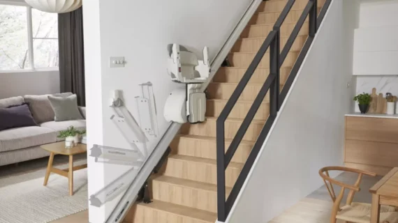 A modern home interior features a wooden staircase with a beige HomeGlide Stair Lift installed along the right side, leading to the second floor. The space also includes a cozy living area and an inviting kitchen.