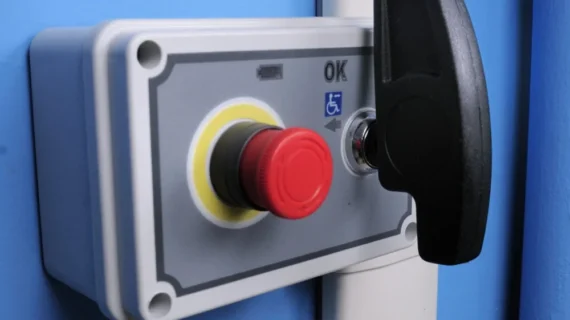 Close-up of a Supra Linea emergency stop button with a red push button, key switch, and OK label. The panel, featuring a wheelchair accessibility symbol, is mounted on a blue wall.