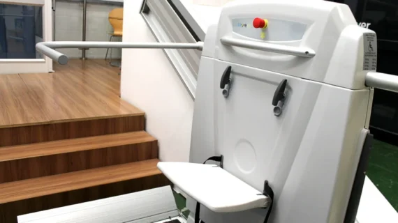 A Supra Linea stair lift with a foldable seat and armrests is installed next to a set of wooden stairs, providing accessibility for people with mobility challenges.