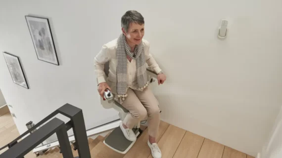 An older woman with short gray hair rides a HomeGlide Stair Lift up a wooden staircase in a modern home, smiling and wearing light-colored clothing and a checkered scarf.
