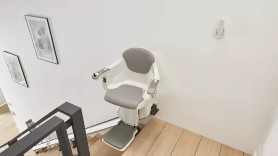 A HomeGlide Stair Lift with a gray and white seat is installed along a wooden staircase in a modern, white-walled home, with framed pictures hanging on the wall.