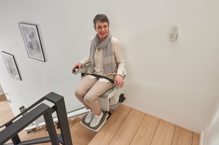 A woman with short gray hair, wearing a scarf and light-colored clothes, sits on a HomeGlide Stair Lift at the top of wooden stairs, smiling and holding the armrest controls.
