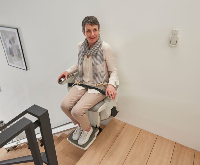 A woman with short gray hair, wearing a scarf and light-colored clothes, sits on a HomeGlide Stair Lift at the top of wooden stairs, smiling and holding the armrest controls.