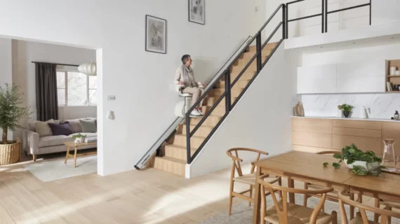 A person uses a HomeGlide Stair Lift to ascend wooden stairs in a modern, open-plan home with a kitchen, dining area, and bright living room.