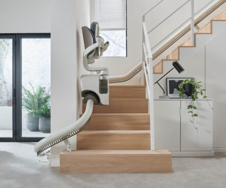 A modern indoor staircase with a light wood finish features a Flow X Curved Stair Lift. Nearby, there’s a white cabinet with a lamp, a plant, and a laptop; large windows and indoor plants provide natural light.