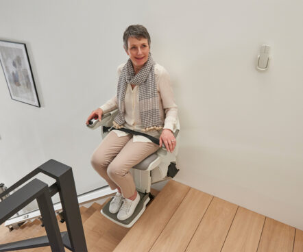 A woman with short gray hair, wearing a scarf and light-colored clothes, sits on a HomeGlide Stair Lift at the top of wooden stairs, smiling and holding the armrest controls.