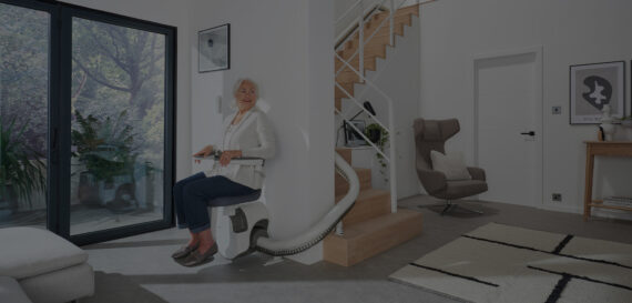 An older woman sits on a stairlift at the base of a wooden staircase in a modern, well-lit living room with large windows, indoor plants, and contemporary furnishings.