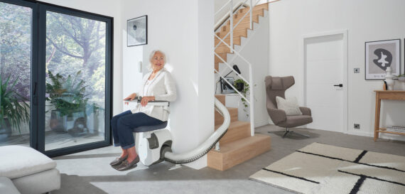 An older woman with gray hair sits on a stairlift at the base of a modern wooden staircase in a bright, stylish living room with large windows and contemporary decor.