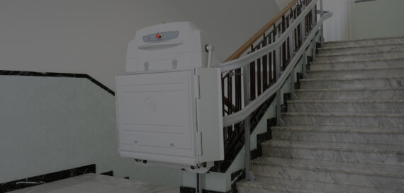 A stair lift platform is attached to a staircase with a marble finish and a wooden handrail, allowing for wheelchair access between floors. The lift is folded up against the side of the stairs.