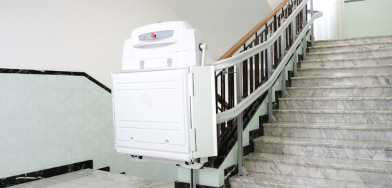 A white stairlift platform is attached to the staircase railing, designed for wheelchair accessibility. The staircase is made of grey marble with black accents and a wooden handrail.
