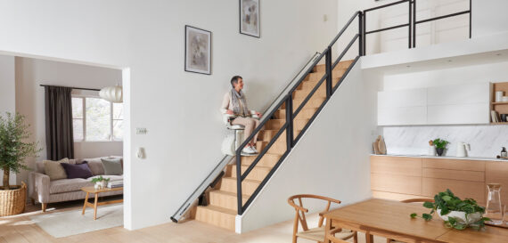 An older adult rides a stairlift up a staircase in a bright, modern home with wooden furniture and white walls. The kitchen, dining, and living areas are visible, decorated with plants and framed pictures.