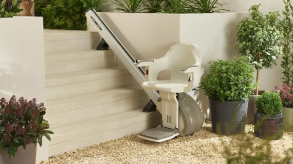 A stair lift chair is installed along the side of outdoor steps, surrounded by green plants and shrubs in a garden setting. The chair, ready for use at the base of the stairs, showcases accessibility solutions similar to those offered by Stair Lifts Chicago.