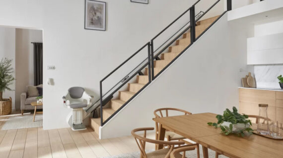 Modern, sunlit interior with a wooden staircase featuring a Stair Lifts Chicago stairlift, a dining table with chairs, light wood floors, framed art on white walls, and a glimpse of a cozy living room with a plant.