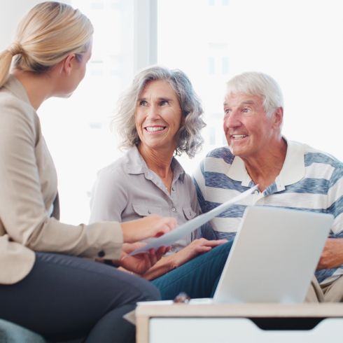 A smiling elderly couple sits on a couch talking with a professional woman holding documents, with a laptop on the table in front of them. The friendly, consultative atmosphere reflects the caring service offered by Windy City Stairlifts - Homepage.