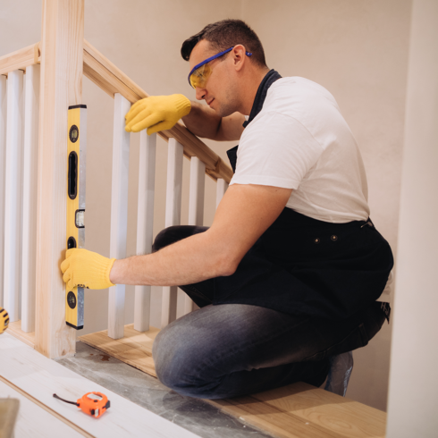 A man wearing yellow gloves and safety glasses uses a spirit level to check the vertical alignment of a wooden stair railing, kneeling while making adjustments—just like the experts at Windy City Stairlifts - Homepage.