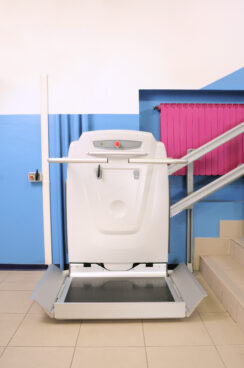 A Platform Lifts motorized platform lift for wheelchairs is installed alongside a staircase with blue walls and a pink radiator in the background, providing accessibility for people with mobility challenges.