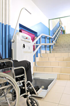A person in a wheelchair prepares to use a platform lift next to a staircase in a brightly lit building, highlighting how platform lifts improve accessibility for individuals with mobility challenges.