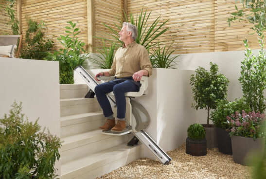 An older man sits on a modern outdoor stairlift on a patio, surrounded by plants and wooden fencing.