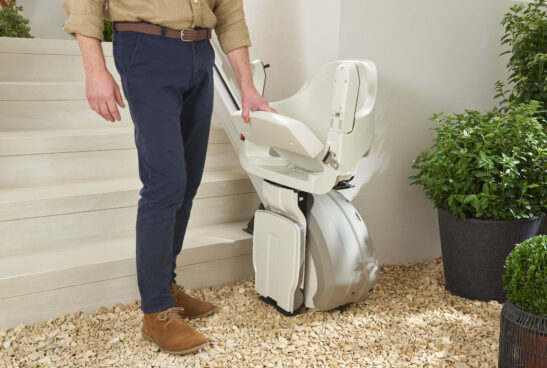 A person stands next to a folded stairlift at the base of indoor stairs, surrounded by potted green plants and light-colored gravel flooring.