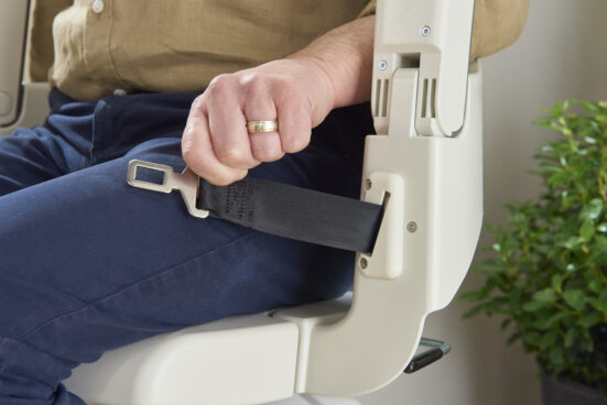 A person wearing blue pants and a beige shirt fastens a black seatbelt on a stairlift chair. A green leafy plant is visible in the background.